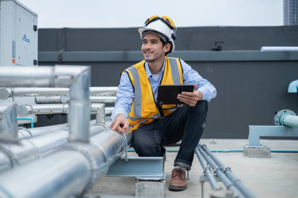 Engineer wearing high-visibility vest and helmet inspects industrial plant, holding a toolbox and tablet for maintenance tasks.